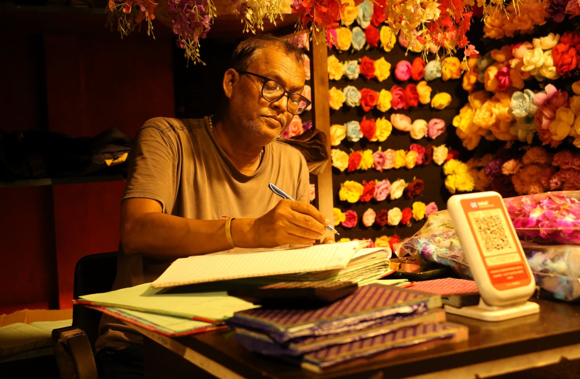 Man surrounded by colorful flowers, writing at desk under soft, warm lighting.